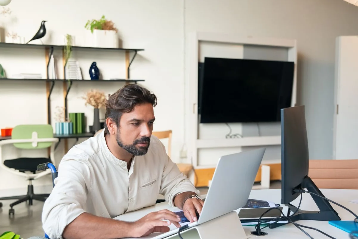 Hombre trabajando con un portátil y un monitor en una oficina luminosa