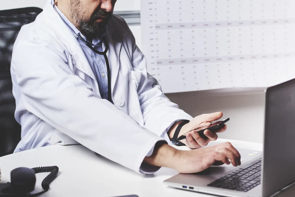 Médico usando el móvil y el portátil en su mesa de consulta