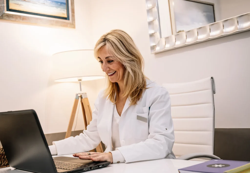 Doctora sonriente trabajando con su portátil en la mesa de la consulta