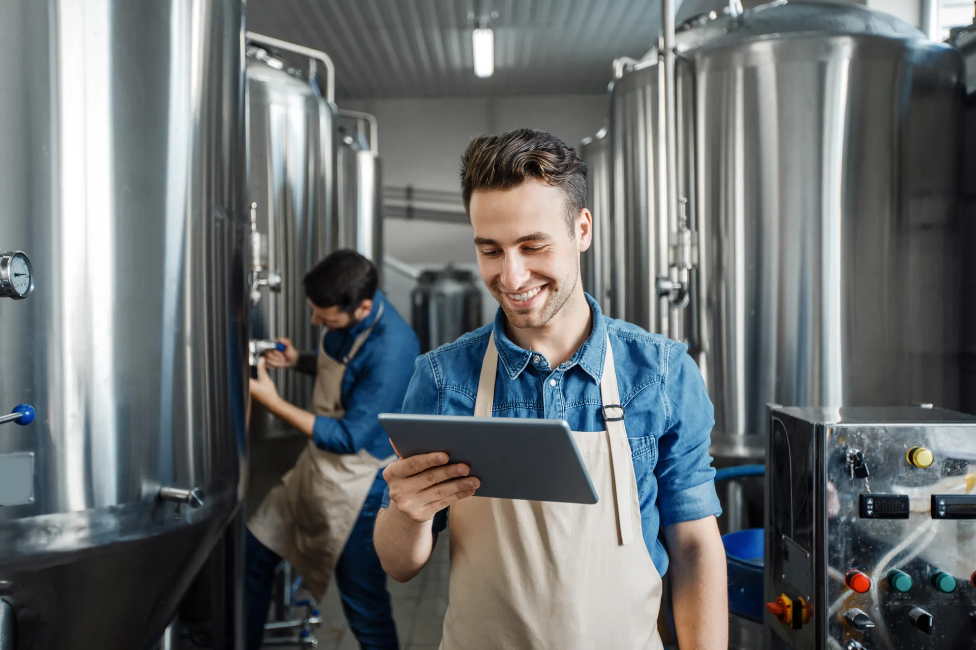 Responsable de una pequeña cervecera consultando la tableta con un empleado trabajando al fondo